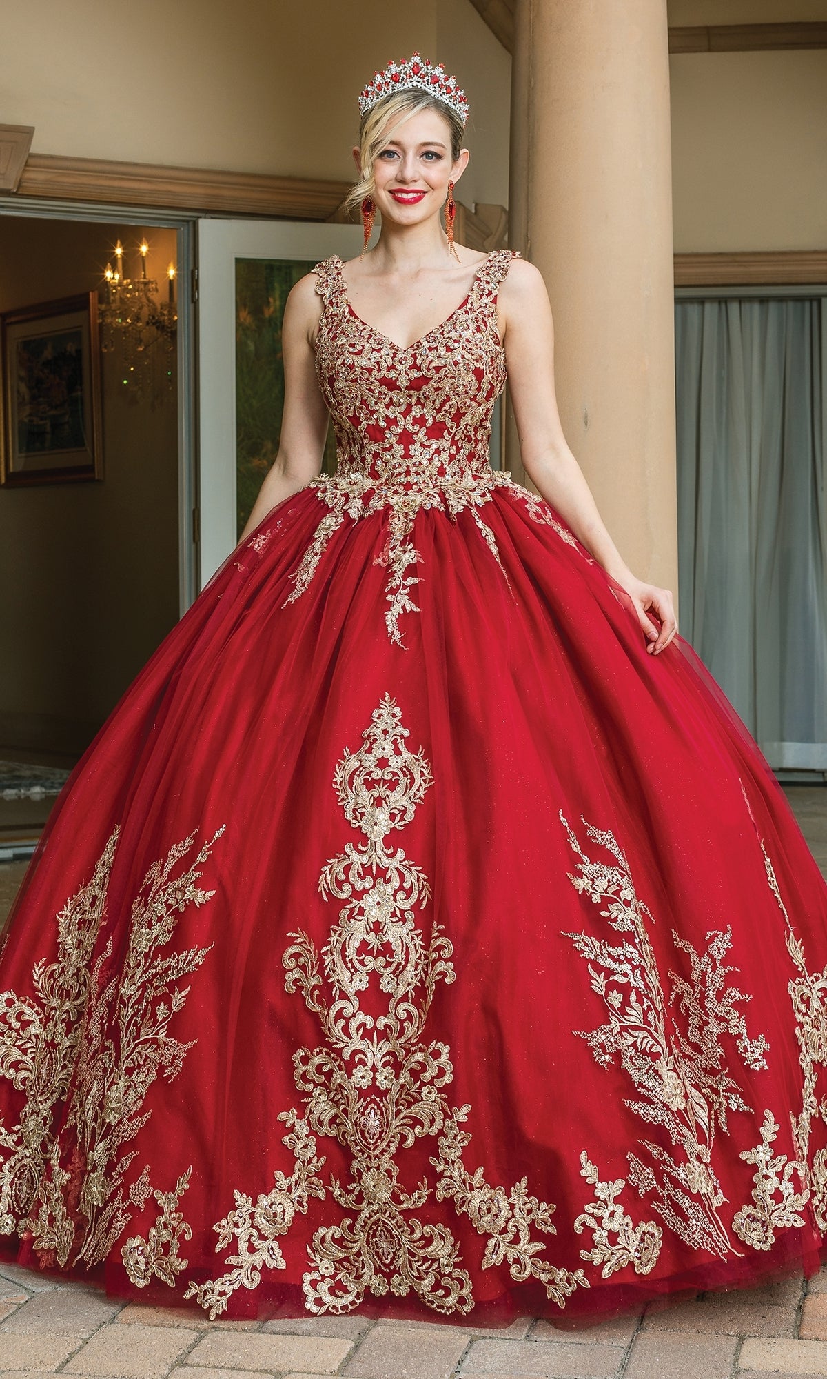 A person stands outside near large columns, wearing the Quinceanera Dress 1612 By Dancing Queen—a burgundy ball gown with gold lace embroidery and a jeweled tiara.